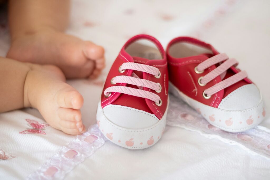 Nackte Babyfüße neben einem Paar roter Stoff-Babyschuhe mit rosa Schnürsenkeln auf weißer Bettwäsche