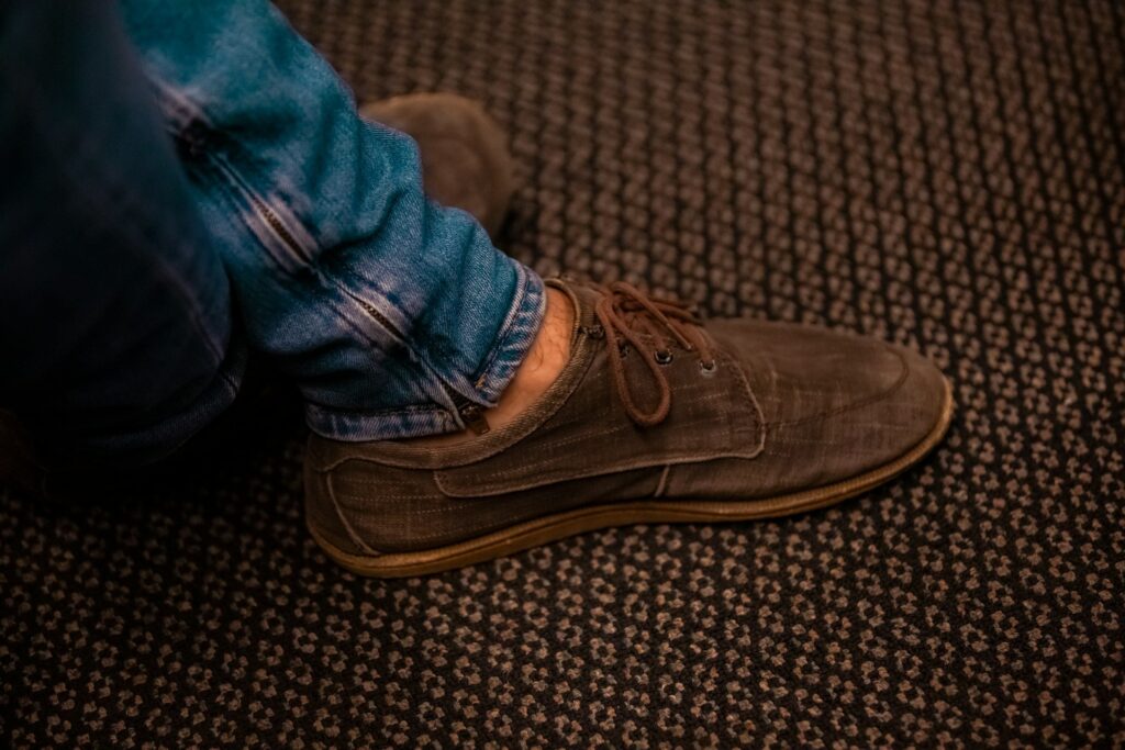 Flacher brauner Stoffschnürschuh mit dünner Sohle zu Jeans auf gemustertem Teppich