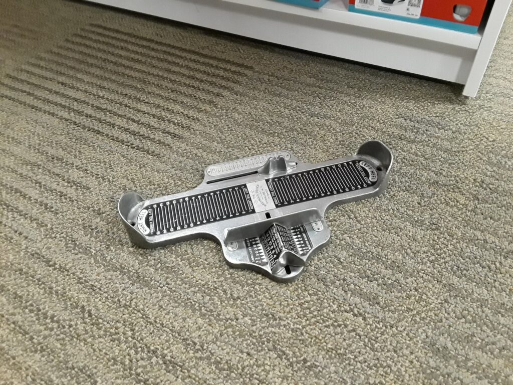 Metallenes Brannock-Messgerät auf grauem Teppich im Schuhgeschäft, oben ein Regal