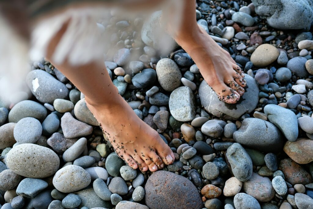 Nackte Füße mit Sand an den Zehen auf grauen Kieseln, ein Fuß unscharf im Schritt