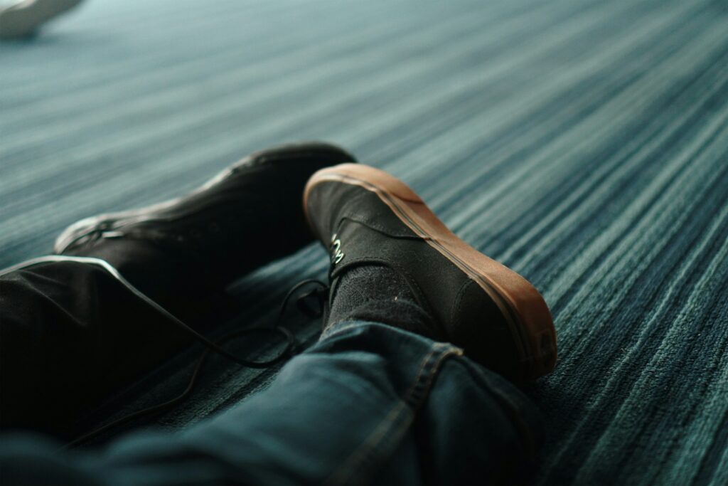 Schwarze Schnürschuhe mit Gummisohle zu Jeans, Beine übereinander auf blau gestreiftem Teppich
