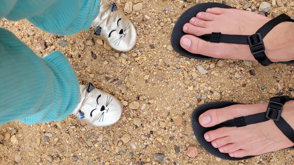 Von oben: Kinderfüße in silbernen Schuhen mit Katzengesicht und Erwachsenenfüße in schwarzen Barfußsandalen auf Kiesboden