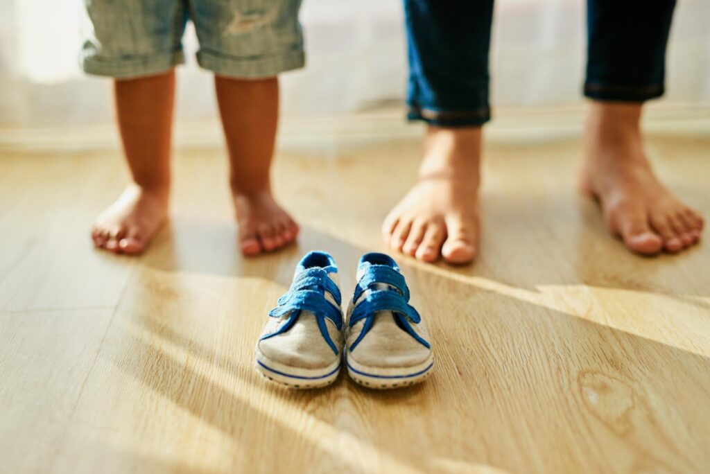 Barfüßige Kinderfüße und Erwachsenenfüße auf Holzboden, davor ein Paar blaue Kinderschuhe