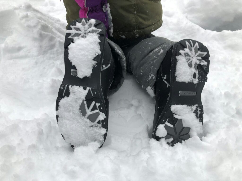 Kind sitzt im Schnee, Blick auf die Sohlen der Winterstiefel mit Schneeflockenprofil