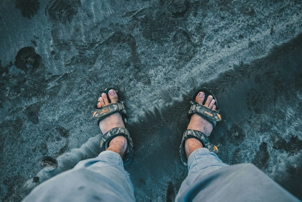 Draufsicht: Füße in schwarzen Wandersandalen mit Tarnmuster-Riemen auf nassem dunklem Sand, Jeans