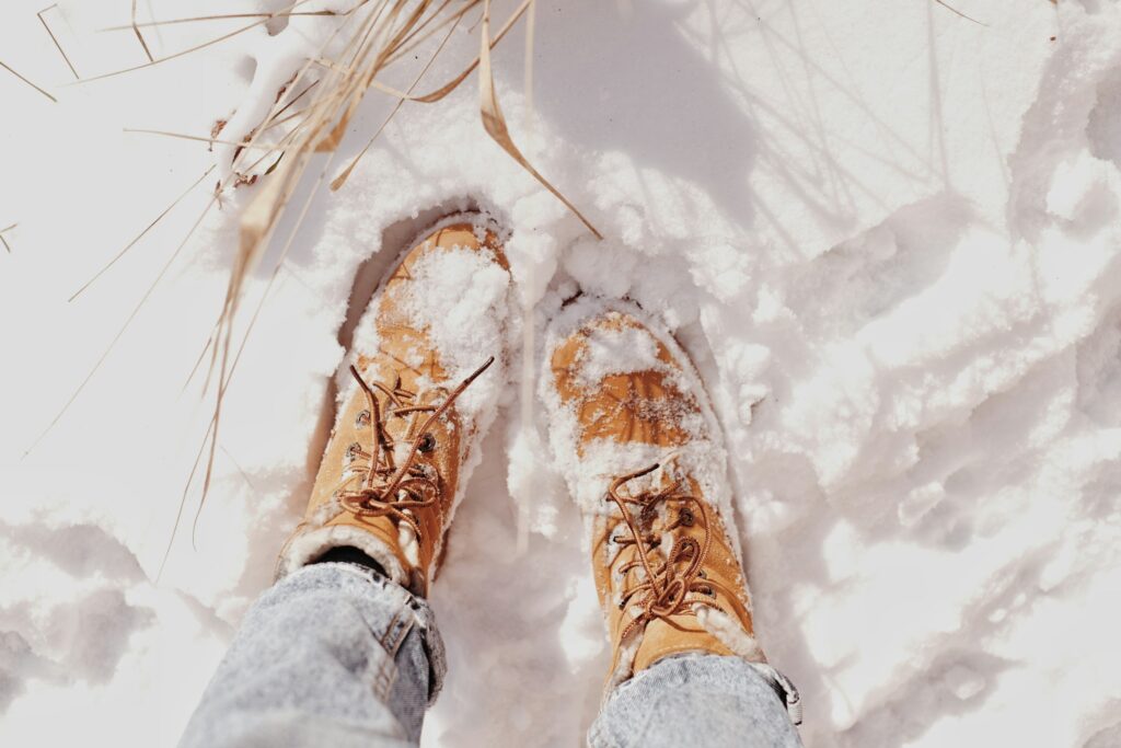 Draufsicht: hellbraune Schnürstiefel mit Schnee auf einem Schneefeld, helle Jeans, trockene Gräser